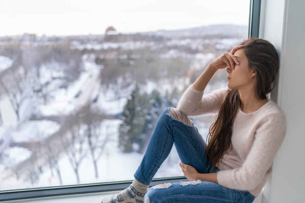 sad woman sitting next to window with cloudy, snowy landscape